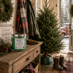 Load image into Gallery viewer, A green scented hand poured candle in a glass jar with a silver lid sits on a rustic wooden table in a cozy entryway. A Christmas tree, winter boots, a plaid scarf, gloves, wreath, and sign are arranged around it. Candle label reads Christmas Tree by Southern Charm Farmhouse.