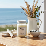 Load image into Gallery viewer, 16 oz hand poured Candle labeled 'Coastal Cottage' with a vase of flowers and pebbles on a table by the ocean. Southern Charm Farmhouse.