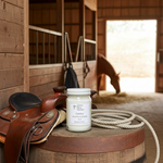 Load image into Gallery viewer, Cowboy Candle in a 16 oz Mason jar on a barrel with a saddle, lasso and a horse in the background inside the stables. Southern Charm Farmhouse.