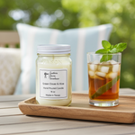 Load image into Gallery viewer, 16 oz mason Jar candle labeled 'Green Clover & Aloe' next to a glass of iced tea with mint leaves on a wooden tray outdoors. Southern Charm Farmhouse