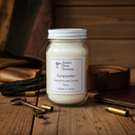 Load image into Gallery viewer, A 16 oz Gunpowder hand-poured candle in a glass Mason jar with a white lid. The candle is centered on a rustic wooden table, surrounded by vintage brass keys, spent shell casings, a dark leather belt, and a leather wallet, set against a warm wood-paneled background. Southern Charm Farmhouse.