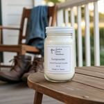 Load image into Gallery viewer, A 16 oz Gunpowder hand-poured candle in a Mason jar with a white lid, sitting on a round wooden porch table. In the blurred background, a pair of leather work boots and a denim jacket draped over a wooden chair sit on a farmhouse-style porch with white railings. Southern Charm Farmhouse.