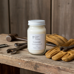 Load image into Gallery viewer, A 16 oz Gunpowder hand-poured candle in a glass Mason jar with a white lid. The candle sits on a weathered wooden workbench alongside vintage tools, including wrenches, a hammer, and a pair of worn leather work gloves, against a rustic wood-paneled wall. Southern Charm Farmhouse.