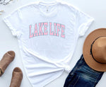 Load image into Gallery viewer, A white t-shirt with 'LAKE LIFE' text in pink gradient, displayed flat with a pair of blue jeans, brown hat, and brown strappy sandals. Southern Charm Farmhouse.
