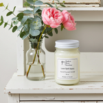 Load image into Gallery viewer, 16 oz mason jar hand poured Love Spell Candle jar with a label next to a vase with pink flowers on a wooden surface. Southern Charm Farmhouse.