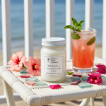 Load image into Gallery viewer, Pink Sands Candle, 16 oz Mason jar on a cocktail on a table with flowers and sea glass, beach in the background. Southern Charm Farmhouse.