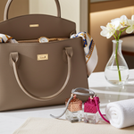 Load image into Gallery viewer, Brown handbag with gold accents, two small car perfume bottles, and a white orchid in a vase on a light surface. Southern Charm Farmhouse.