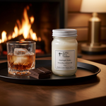 Load image into Gallery viewer, A Southern Charm Farmhouse mason jar candle labeled Midnight Rider sits on a matte black tray next to a crystal lowball glass of bourbon with a large square ice cube and two squares of dark chocolate, set against the warm glow of a roaring fireplace background.