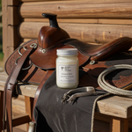 Load image into Gallery viewer, A rustic Wild West product display featuring the Midnight Rider hand-poured candle sitting on black fabric draped over a weathered wooden bench, leaning against a detailed brown leather horse saddle with silver spurs and a lasso rope in the foreground. Southern Charm Farmhouse.