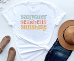 Load image into Gallery viewer, A white t-shirt with the text 'Saltwater, Seashells, Sunshine' printed in colorful letters, displayed flat with a pair of blue jeans, a pair of brown sandals, and a straw hat. Southern Charm Farmhouse.