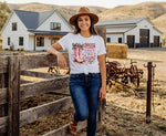 Load image into Gallery viewer, Woman standing in a barnyard wearing a white t-shirt with graphic design and blue jeans, with a farmhouse in the background. Southern Charm Farmhouse.