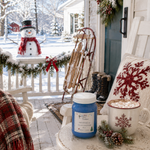 Load image into Gallery viewer, A cozy winter porch scene with a blue hand poured candle labeled Jack Frost in the foreground and a snowy outdoor porch setup behind it, including a snowman decoration, wreath, boots, sled, pillow, and mug of hot chocolate.  Southern Charm Farmhouse.
