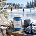 Load image into Gallery viewer, A blue hand poured candle labeled 'Jack Frost' sits on a wooden table outdoors in a snowy winter scene, with ice skates, a mug, gloves, evergreen branches, and a frozen lake in the background. Southern Charm Farmhouse.
