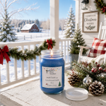 Load image into Gallery viewer, A lit blue hand-poured Jack Frost candle in a clear glass jar sits on a snowy porch decorated for Christmas, with snow-covered trees, a red plaid pillow, pinecones, and a white house background. Southern Charm Farmhouse.
