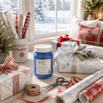 Load image into Gallery viewer, A styled holiday gift-wrapping scene by a window with wrapped presents, gift bags, ribbon spools, wrapping paper rolls, scissors, and a blue hand poured candle labeled Jack Frost in the foreground. The setting includes a window view of snow and pillows on a couch in the background. Southern Charm Farmhouse.
