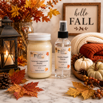 Load image into Gallery viewer, Two fall-themed scented products from Southern Charm Farmhouse: a jar candle labeled 'Caramel Cappuccino Brulee' and a room spray bottle labeled 'Vanilla Maple Pecan.' The products are styled on a marble surface with autumn leaves, pumpkins, a lantern, a knitted blanket, and a framed 'hello FALL' sign in the background.
