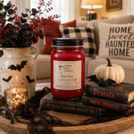 Load image into Gallery viewer, A red hand-poured candle in a 16 oz mason  jar with a black lid and a white label reading 'Bite Me',  styled on a tray with books, a small pumpkin, a candle, and a ceramic vase in a cozy living room setting. Southern Charm Farmhouse.
