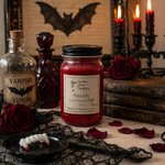 Load image into Gallery viewer, A Halloween-themed tabletop scene featuring a red hand-poured candle in a 16 oz mason  jar with a black lid and a label reading 'Southern Charm Farmhouse Bite Me'. The candle is styled with dark red roses, black lace, a plate with fangs, a bottle of vampire venom, stacked gothic books, and lit candles in the background.
