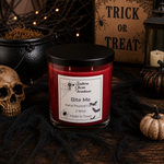 Load image into Gallery viewer, A red scented candle in a clear glass jar with a black lid sits on a dark surface surrounded by Halloween props, including a skull, black webbing, a small pumpkin, a cauldron, and a sign that reads 'Trick or Treat.' The front label shows the brand Southern Charm Farmhouse and the scent/name 'Bite Me.

