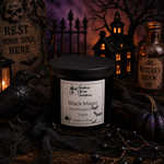 Load image into Gallery viewer, A lit black scented candle in a clear glass jar sits on a rustic wooden surface surrounded by Halloween decor, including a black raven, cobwebbons, a small purple pumpkin, and a bottle labeled 'Witches Brew.' The candle label reads 'Southern Charm Farmhouse' and 'Black Magic Hand Poured Candle 2 Wick Made in Texas.
