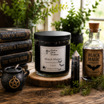 Load image into Gallery viewer, A gothic-themed candle set on a rustic wooden table, featuring a 'Black Magic' hand-poured candle in a glass jar on a wood slice, a black cauldron, a crystal point, and a vintage-style apothecary bottle labeled 'Black Magic Apothecary'.  Southern Charm Farmhouse.
