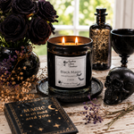 Load image into Gallery viewer, A lit black scented hand poured candle in a clear glass jar sits on a dark coaster on a rustic table, surrounded by gothic props including a vase of black roses, a black skull, a black book, a black goblet, and a glittery bottle. The candle label reads 'Black Magic. Southern Charm Farmhouse.
