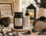 Load image into Gallery viewer, One Southern Charm Farmhouse Hand Poured Candle products, a jar candle labeled 'Black Magic' and a room spray bottle labeled 'Black Magic Room Spray', displayed on a rustic table surrounded by Halloween-themed props including a mug, cookies, spider decorations, and books.
