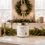 Load image into Gallery viewer, A hand-poured Christmas Cheer candle in a clear glass jar with a silver lid sits on a marble countertop decorated with green pine branches, gold candlesticks, and a lit holiday wreath in the background. Southern Charm Farmhouse.

