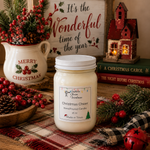 Load image into Gallery viewer, A hand-poured Christmas candle in a clear 16 oz mason jar with a white lid sits on a rustic wooden table surrounded by holiday decorations, including a white pitcher with 'Merry Christmas' text, red berries, pinecones, a plaid cloth, and a small lit decorative house. Candle label reads Christmas Cheer. Southern Charm Farmhouse.
