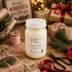 Load image into Gallery viewer, A hand-poured Christmas Cheer candle in a clear 16 oz mason jar with a white lid, surrounded by holiday-themed props like wrapped gifts, pine branches, scissors, and a spool of ribbon on a wooden surface. Southern Charm Farmhous.
