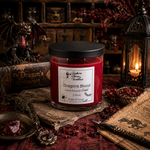 Load image into Gallery viewer, A red hand-poured candle in a clear glass jar with a black lid, labeled 'Dragons Blood' by Southern Charm Farmhouse., styled on an old book surrounded by gothic props including a dragon figurine, lantern, map, and dried red flowers.
