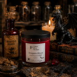 Load image into Gallery viewer, A red hand-poured wax candle in a clear glass jar with a black lid, labeled 'Dragons Blood' by Southern Charm Farmhouse, styled on a dark table with a black dragon figurine, potion bottles, books, and dried herbs.
