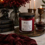 Load image into Gallery viewer, A red Dragon's Blood scent hand poured candle in a clear glass jar with a black lid sits on a round brass tray beside a small glass of matching dark red liquid. The scene is styled with books, a black dragon figurine, a vase of red flowers, and lit candles in a warm indoor setting. Southern Charm Farmhouse

