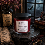 Load image into Gallery viewer, A red wax candle in a clear glass jar with a black lid and a white label reading 'Southern Charm Farmhouse' and 'Dargons Blood', styled on a dark surface with gothic props including a potion bottle, old books, a key, and a dark window background.
