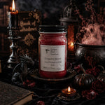 Load image into Gallery viewer, A gothic-themed hand poured candle in a 16 oz mason jar labeled 'Dragons Blood' by Southern Charm Farmhouse., styled with a black lid, surrounded by candles, a cauldron, a dragon figurine, and dark pumpkins on a dark cloth.
