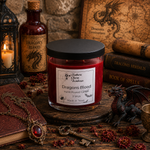 Load image into Gallery viewer, A hand-poured candle in a clear glass jar with a black lid and a white label reading 'Dragons Blood' by Southern Charm Farmhouse, styled on a rustic wooden surface with books, a lantern, and a small dragon figurine.
