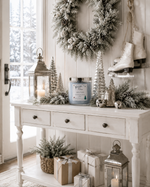 Load image into Gallery viewer, A styled holiday entryway scene with a white console table holding a blue hand poured candle labeled Jack Frost, silver lanterns, frosted greenery, small silver trees, wrapped gifts, a wreath, and ice skates hanging on the wall. Southern Charm Farmhouse
