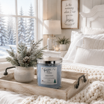 Load image into Gallery viewer, A hand-poured Jack Frost candle in a clear glass jar with a silver lid sits on a rustic wooden tray on a cozy bed. The room is styled with white bedding, a small potted pine branch, and a window showing a snowy outdoor view. Southern Charm Farmhouse
