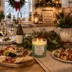 Load image into Gallery viewer, A lit green scented hand poured candle in a clear glass jar sits on a kitchen table surrounded by holiday food and decor, including a bottle of wine, charcuterie boards, grapes, crackers, and festive greenery. The candle label reads 'Mistletoe'. Southern Charm Farmhouse.

