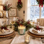 Load image into Gallery viewer, A lit scented hand poured candle in a clear glass jar sits on a rustic holiday table setting, surrounded by white wine glasses, plates with folded napkins tied with red ribbon, woven placemats, and evergreen garlands with red berries. The candle label reads 'Mistletoe' and shows a green wax color. Southern Charm Farmhouse.
