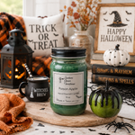 Load image into Gallery viewer, A green wax hand poured candle in a 16 oz mason jar with a black lid and a white label reading 'Poison Apple' by Southern Charm Farmhouse, styled on a wooden board with Halloween props including a lantern, mug, pumpkin, books, and a sign.
