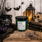 Load image into Gallery viewer, A Halloween-themed hand poured candle labeled 'Poison Apple' sits on a rustic wooden table surrounded by spooky props including a cauldron, lantern, skull, books, dried lavender, and black lace against a white wood plank background. Southern Charm Farmhouse.
