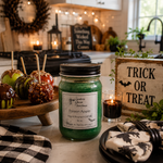 Load image into Gallery viewer, A hand-poured green candle in a 16 oz mason jar with a black lid and a white label reading 'Poison Apple' by Southern Charm Farmhouse, styled on a kitchen counter with Halloween decorations, candy apples, and a 'Trick or Treat' sign.
