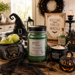 Load image into Gallery viewer, A Halloween-themed tabletop scene featuring a green hand-poured candle in a 16 oz  mason jar labeled 'Poison Apple' by Southern Charm Farmhouse. The jar is surrounded by witch hats, a black bowl of green apples, a small 'Poison Apple' sign, a black mug reading 'Hocus * Pocus' with whipped cream and a striped straw, a lit jack-o'-lantern, cobwebs, and a rustic sign that says 'Welcome to Foolish Mortals'.
