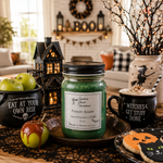 Load image into Gallery viewer, A Halloween-themed hand poured candle in a 16 oz mason jar with a black lid and a white label reading  'Poison Apple' Hand Poured Candle 16 oz Made in Texas with love' sits on a wooden tray. The scene is styled with a black cauldron of green apples, a small haunted house lantern, a gory apple, and a black mug that says 'WITCHES GET STUFF DONE'. Southern Charm Farmhouse.
