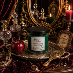 Load image into Gallery viewer, A hand-poured scented Poison Apple candle in a green glass jar with a black lid sits on an ornate silver tray. The candle is styled with a red apple, crystal perfume bottles, a gold mirror, a lit purple candle in a brass holder, and a small sign reading 'Fairiest of All. Southern Charm Farmhouse.
