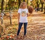 Load image into Gallery viewer, Woman in a white t-shirt with a pumpkin design, holding a brown hat, standing in an autumn park. Southern Charm Farmhouse.
