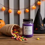 Load image into Gallery viewer, A purple hand-poured candle in a glass jar with a black lid and a white label reading 'Trick or Treat' by Southern Charm Farmhouse, styled on a wooden table with candy, a burlap sack, a witch hat, and orange string lights in the background.
