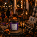 Load image into Gallery viewer, A Halloween-themed candle jar with a purple label reading 'Trick or Treat' sits on a round table in a decorated porch setting at night. The scene includes a lantern, a plaid chair with a 'Trick or Treat' pillow, candy bowls, pumpkins, and a 'Welcome' sign in the background. Southern Charm Farmhouse.
