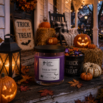 Load image into Gallery viewer, A purple scented hand poured candle labeled Trick or Treat in a clear glass jar with a black lid sits on a rustic porch surrounded by Halloween decor, including pumpkins, a lantern, a bucket of candy, fall leaves, and a porch chair at dusk. Southern Charm Farmhouse.
